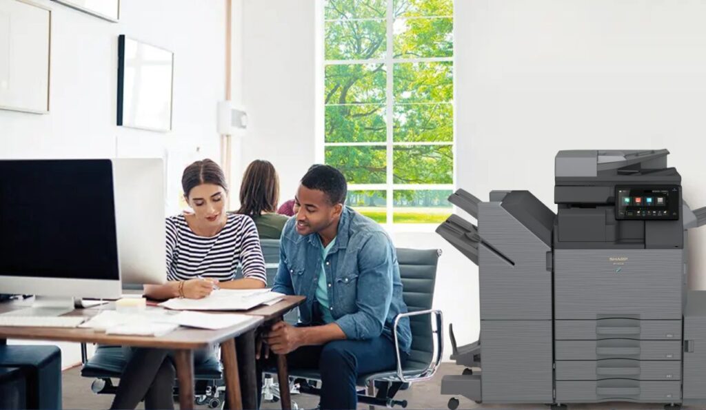 Oficina moderna con empleados trabajando en equipo junto a una impresora multifunción Sharp BP-50C36 A3 Color.